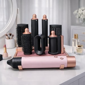 Pode incluir: Um conjunto de ferramentas de modelagem de cabelo, incluindo um secador de cabelo rosa e ouro rosa e vários acessórios pretos. As ferramentas estão dispostas em uma penteadeira com um espelho e outros produtos de beleza. A estética geral é moderna e elegante.
