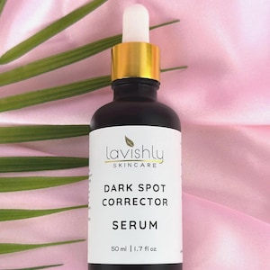 May include: A black skincare serum bottle with a gold cap and white dropper. The label reads "Lavishly Skincare" and "Dark Spot Corrector Serum". The bottle contains 1.7 fl oz of serum. The background is pink with green leaves.