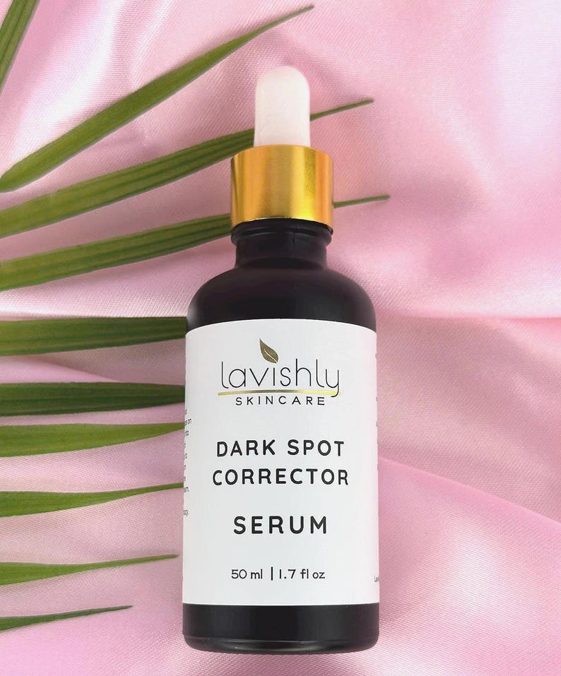 May include: A black skincare serum bottle with a gold cap and white dropper. The label reads "Lavishly Skincare" and "Dark Spot Corrector Serum". The bottle contains 1.7 fl oz of serum. The background is pink with green leaves.