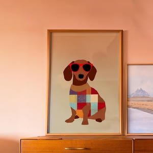 Peut inclure: Impression encadrée d'un teckel marron portant des lunettes de soleil rouges et un pull patchwork coloré. Le chien est assis, sur un fond beige clair. L'impression est exposée sur une commode en bois.