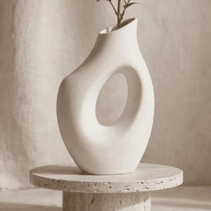 Könnte beinhalten: Eine matte, cremeweiße Keramikvase mit asymmetrischer, organischer Form und einem großen, hohlen, ovalen Ausschnitt in der Mitte. Die Vase hat eine schmale, schräge Öffnung an der Oberseite und eine glatte, durchgehend solide Oberflächenstruktur.