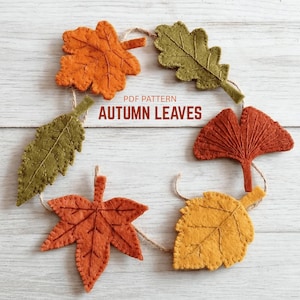 May include: A garland of six felt autumn leaves in orange, yellow, and olive green shades, connected by a thin brown twine. The leaves feature stitched vein details. Text in the center reads: PDF PATTERN AUTUMN LEAVES.