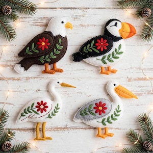 Op de afbeelding: Vier viltvogelornamenten met geborduurde rode bloemen en groene bladeren. De set bevat een bruine en witte adelaar, een zwart-witte papegaaiduiker met een oranje snavel, een witte ooievaar en een grijze en witte pelikaan.
