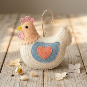 May include: A felt chicken ornament featuring a cream-colored body, a peach-colored head with a yellow beak, and a blue wing patch with a peach heart. The ornament includes a brown twine loop for hanging and decorative stitching throughout.