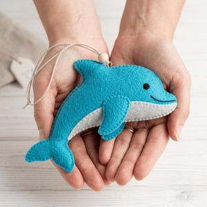 Op de afbeelding: Een vilten dolfijnornament met een fel turquoise blauwe bovenkant en een lichtgrijze onderbuik. De dolfijn heeft een klein zwart geborduurd oog en een gebogen mond. Een dun beige touwtje is aan de bovenkant bevestigd met een houten kraal.