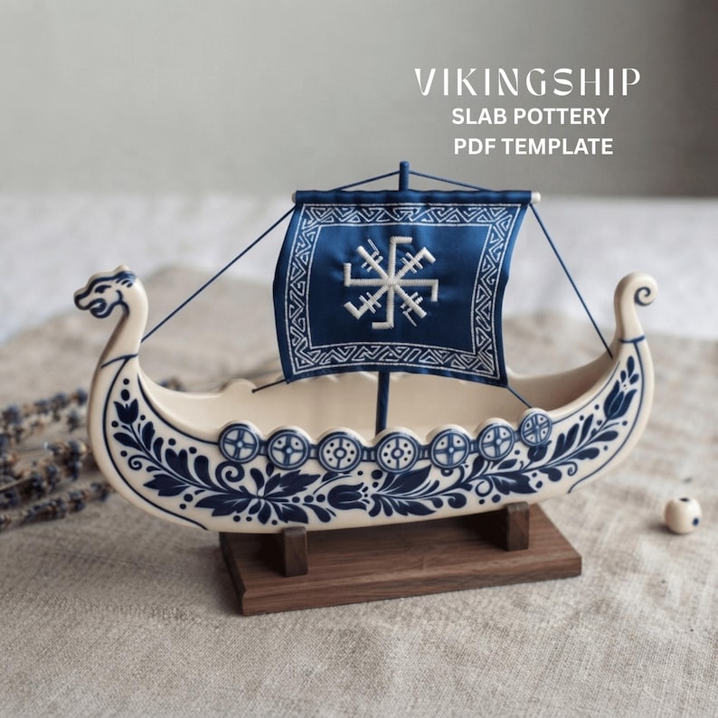 May include: A ceramic Viking ship model featuring cream-colored pottery with intricate blue floral patterns and circular shield designs. It includes a dark blue fabric sail with a white geometric symbol and the text VIKINGSHIP SLAB POTTERY PDF TEMPLATE.