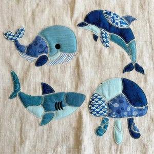 May include: Four fabric appliques stitched onto a beige linen background. The designs include a blue whale, a leaping dolphin, a shark with gills, and a jellyfish, all crafted from various shades of blue patterned cotton fabric.
