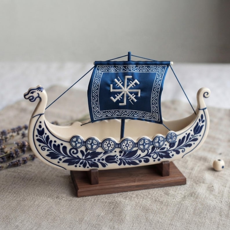 May include: A ceramic model of a Viking ship featuring a cream-colored hull decorated with blue floral patterns and circular shield motifs. The ship has a blue fabric sail embroidered with a white geometric sun symbol, mounted on a wooden base.