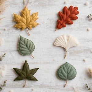 May include: A set of six felt leaf ornaments in various shapes and colors. The collection includes a yellow maple leaf, a rust-colored oak leaf, a sage green leaf, a cream ginkgo leaf, a dark olive star-shaped leaf, and a round green leaf.