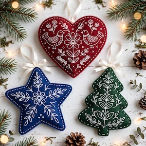 Puede incluir: Tres adornos de fieltro con patrones bordados en blanco: un corazón rojo con pájaros y flores, una estrella azul con un motivo floral central y un árbol de Navidad verde con diseños de hojas. Cada adorno tiene una cinta blanca para colgar.