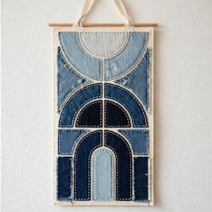 May include: A rectangular wall hanging made of layered denim fabric patches in various shades of blue, ranging from light wash to dark indigo. The textile art features geometric shapes including arches and triangles, held by a wooden rod.