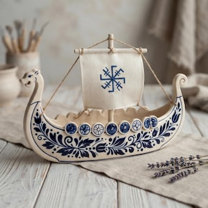 May include: A ceramic model of a Viking ship featuring a cream-colored hull decorated with blue floral patterns and a row of circular shields. The ship has a single mast with a rectangular off-white fabric sail displaying a blue geometric symbol.