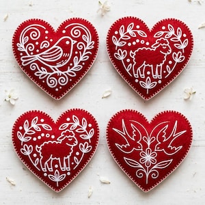 May include: Four heart-shaped felt ornaments in deep red with white embroidered outlines. The designs feature a bird, two sheep, and two birds facing a central flower, all surrounded by delicate white leaf and floral patterns with stitched edges.