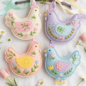 May include: Four felt hen ornaments in pastel colors: white, lavender, peach, and blue. Each hen has floral and leaf embroidery, with details like pink accents, yellow beaks, and small ribbons for hanging. The ornaments are displayed on a white surface.