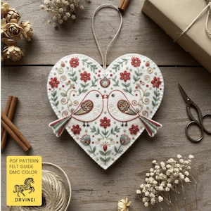 May include: A heart-shaped felt ornament with two kissing birds, red flowers, and green leaves. The ornament has a cream background and a string for hanging. The image also includes a spool of twine, scissors, and a wrapped gift.