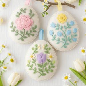 May include: Three felt Easter egg ornaments with floral designs. One features a pink flower, another blue flowers, and the third a purple tulip. Each egg has a ribbon for hanging. Daisies and tulips surround the eggs.