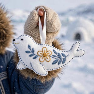 Può includere: Un ornamento in feltro bianco a forma di foca con motivi a foglie ricamati in blu e un fiore giallo centrale. La foca è fissata a un nastro bianco per essere appesa, con cuciture visibili lungo i bordi del corpo in feltro.