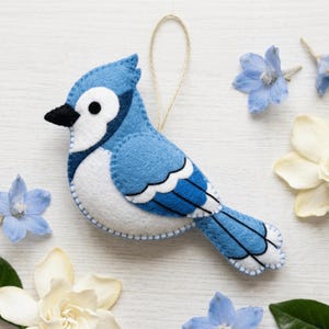 May include: A felt blue jay ornament featuring shades of blue, white, and black with visible stitching along the edges. The bird has a pointed black beak, a round black eye, and a thin tan string loop attached to the top for hanging purposes.