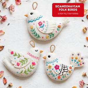 May include: Three white felt bird ornaments with embroidered floral patterns in pink, blue, yellow, and green. Each bird has a small jute loop for hanging. A red rectangular graphic in the corner displays the text SCANDINAVIAN FOLK BIRDS 3 PDF FELT PATTERNS.