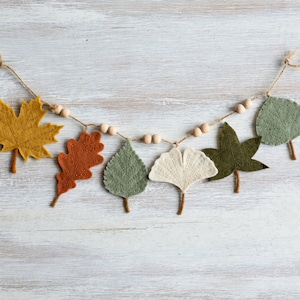 May include: A garland featuring six felt leaves in shades of mustard yellow, burnt orange, sage green, cream, and olive green, interspersed with small, round, natural wooden beads strung along a thin, light brown twine cord for hanging.