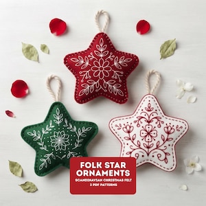 May include: Three star-shaped felt ornaments with white hanging loops. One is deep red with white floral embroidery, one is dark green with white floral embroidery, and one is white with red floral embroidery. Text reads: FOLK STAR ORNAMENTS.