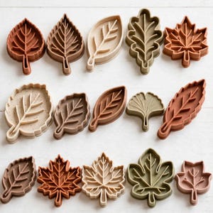 Peut inclure: Une collection de quinze emporte-pièces en forme de feuilles disposés sur trois rangées. Les emporte-pièces sont déclinés dans diverses nuances de brun, de beige, de crème et de vert doux, avec des nervures détaillées moulées au centre.