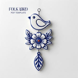 Op de afbeelding: Een hangend ornament met een witte vogel met blauwe omlijning, een centrale bloem met blauwe bloemblaadjes en een rood hart, en een blauw-wit blad onderaan. De tekst FOLK BIRD PDF TEMPLATE is zichtbaar in de linkerbovenhoek.
