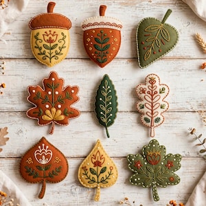 May include: A collection of nine felt ornaments shaped like autumn leaves and acorns, featuring intricate embroidery with floral and botanical patterns in shades of orange, yellow, green, and white, arranged in a grid on a light wood surface.