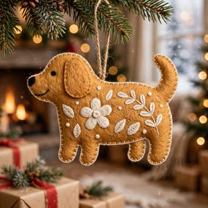 Puede incluir: Un adorno de fieltro en forma de perro de color marrón dorado con bordados blancos de flores y hojas. La figura del perro tiene los bordes cosidos y cuelga de un cordel de yute fino. El diseño incluye una flor central.