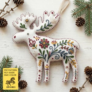May include: A white felt moose decoration with colourful embroidered floral patterns. The ornament features a cream-coloured hanging loop and is surrounded by pine cones and evergreen branches. The text "PDF PATTERN FELT GUIDE DMC COLOR" is visible.