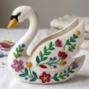 Op de afbeelding: Een handgemaakte witte vilten zwaan met geborduurde bloemen. De zwaan heeft een gele snavel, zwarte ogen en vleugels versierd met paarse, rode en gele bloemen en groene bladeren. Een decoratief item voor woondecoratie.