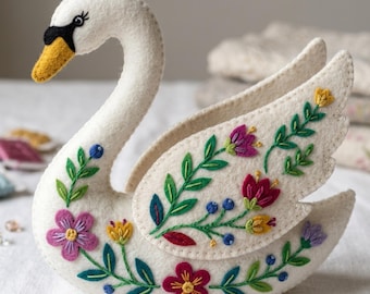 Peut inclure: Un cygne en feutre blanc fait à la main, orné de broderies florales. Le cygne a un bec jaune, des yeux noirs et des ailes ornées de fleurs violettes, rouges et jaunes, et de feuilles vertes. Un objet décoratif pour la décoration intérieure.