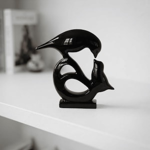 May include: A glossy black ceramic sculpture featuring two stylized birds facing each other. The upper bird is positioned above the lower bird, creating a continuous, fluid, and curved silhouette. The piece stands on a rectangular base.