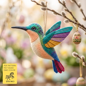 May include: A felt hummingbird ornament featuring a teal head, peach chest, and multi-coloured wings in purple, teal, and yellow. The bird has a black beak and a red and blue tail. A yellow square label displays the text PDF PATTERN FELT GUIDE DMC COLOR.