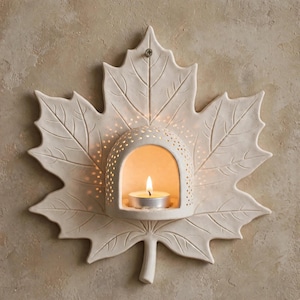 May include: A cream-colored ceramic wall sconce shaped like a maple leaf with a central dome featuring perforated holes. A small lit tea light candle sits inside the dome, casting a warm glow through the openings in the ceramic structure.