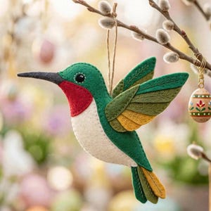May include: A felt hummingbird ornament featuring a dark green back, a bright red throat patch, and a cream-colored belly. The wings are layered in shades of olive and mustard green with stitched details. It hangs from a thin brown twine loop.