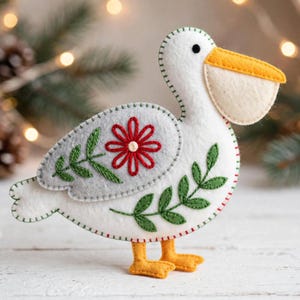 May include: A felt pelican ornament featuring a white body, a yellow beak, and orange feet. The wing is light gray with a red embroidered flower and green leaf stitching. The body is decorated with green embroidered leaves and green edge stitching.