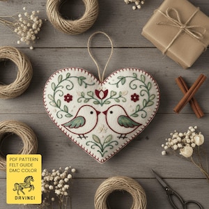 May include: A heart-shaped felt ornament featuring hand-embroidered details of two birds facing each other, surrounded by green vines, red flowers, and small white accents. The ornament includes a jute hanging loop at the top centre.
