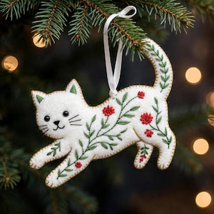 May include: A white felt cat ornament featuring embroidered green vines and small red flowers. The cat has black stitched eyes, a nose, and whiskers. A white ribbon loop is attached to the top for hanging, with visible stitching along the edges.
