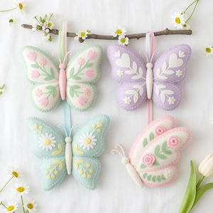 May include: Four felt butterfly decorations in pastel shades, each featuring floral embroidery. The butterflies are white, light green, light blue, and lavender, with pink, yellow, and white floral details. They are suspended from a small branch with white flowers.