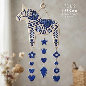Puede incluir: Un adorno de cerámica color crema con forma de caballo y patrones de arte popular azul, con tres cuerdas colgantes de formas azules que incluyen estrellas, corazones, flores y hojas. El texto en la imagen dice FOLK HORSE 12 STRING ELEMENTS PDF - SVG.