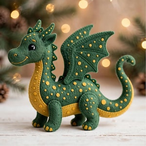 Puede incluir: Una figura de dragón de fieltro hecha a mano en verde oscuro con vientre amarillo y lunares amarillos. La criatura presenta alas cosidas, una cola curva y una expresión sonriente con grandes ojos negros, apoyada sobre cuatro patas firmes.