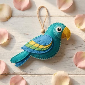 May include: A felt parrot ornament featuring a teal body, a yellow and green layered wing, and a yellow beak. The bird has a white and black eye, is stitched with white thread, and includes a tan wooden bead attached to a jute hanging loop.
