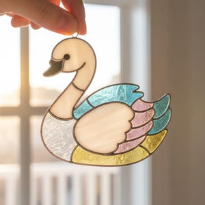 May include: A stained glass suncatcher shaped like a swan with a cream-coloured neck and head, a textured white body, and wings featuring segments of iridescent light blue, soft pink, and pale yellow glass held together by dark metal solder lines.