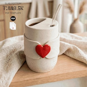 May include: A beige speckled ceramic tumbler with a lid featuring a straw hole and a clear glass straw. A bright red heart is attached to the center of the tumbler. Text in the image reads SLAB POTTERY TUMBLER PDF TEMPLATE and PDF.