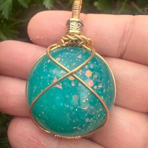 May include: A teal-colored pendant with glitter and gold wire wrapping. The round, glittery orb is encased in a gold wire cage, with a gold wire loop at the top for hanging. The pendant is held in a hand against a background of green foliage.