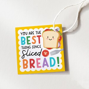 May include: A square yellow gift tag with a white scalloped border and a white string. The text reads YOU ARE THE BEST THING SINCE SLICED BREAD! It features a cartoon illustration of a smiling slice of bread, a tomato slice, and a silver fork.
