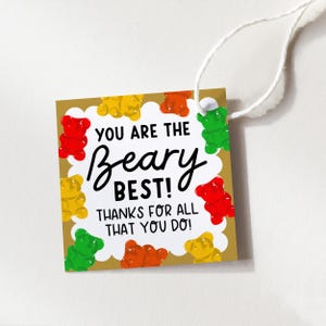 May include: A square gift tag with a gold border and white cloud-like accents. The tag features colorful gummy bear illustrations and the text "YOU ARE THE Beary BEST! THANKS FOR ALL THAT YOU DO!" A white string is attached.