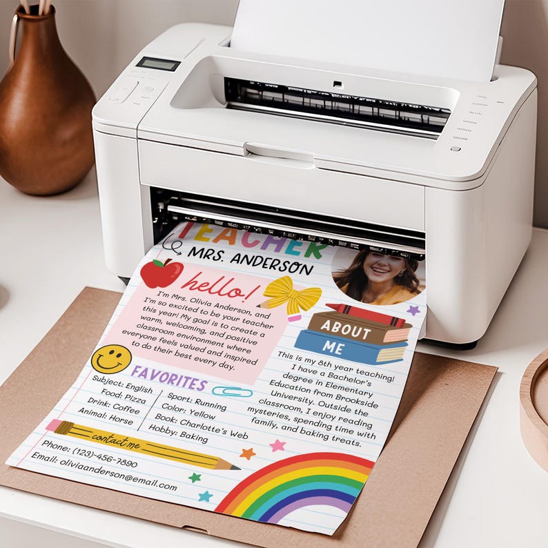 May include: A white desktop printer outputting a printed teacher introduction sheet. The document features the text 'TEACHER MRS. ANDERSON', a photo of a person, a red apple, a yellow bow, a stack of books, a smiley face, and a rainbow graphic.