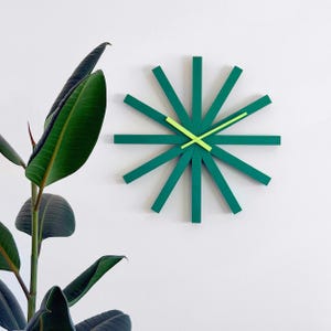 Puede incluir: Un reloj de pared moderno con un diseño de estrella. El reloj presenta brazos rectangulares de color verde oscuro que irradian desde el centro, con manecillas de color verde neón brillante. El reloj está montado en una pared blanca, con una gran planta verde en primer plano.